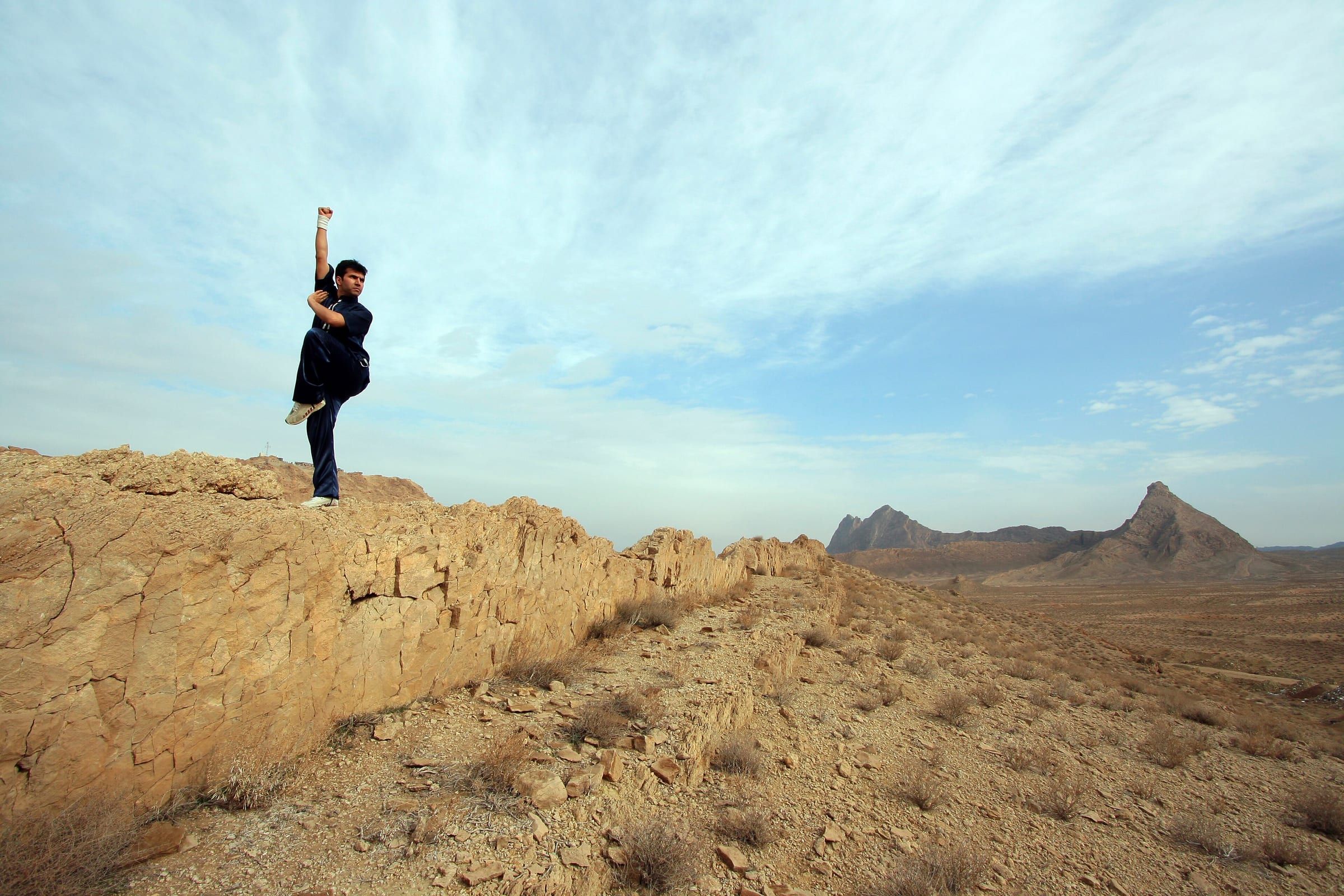 Kung-Fu-Sportler in hohem Einbeinstand auf einem Felsen unter großem Himmel