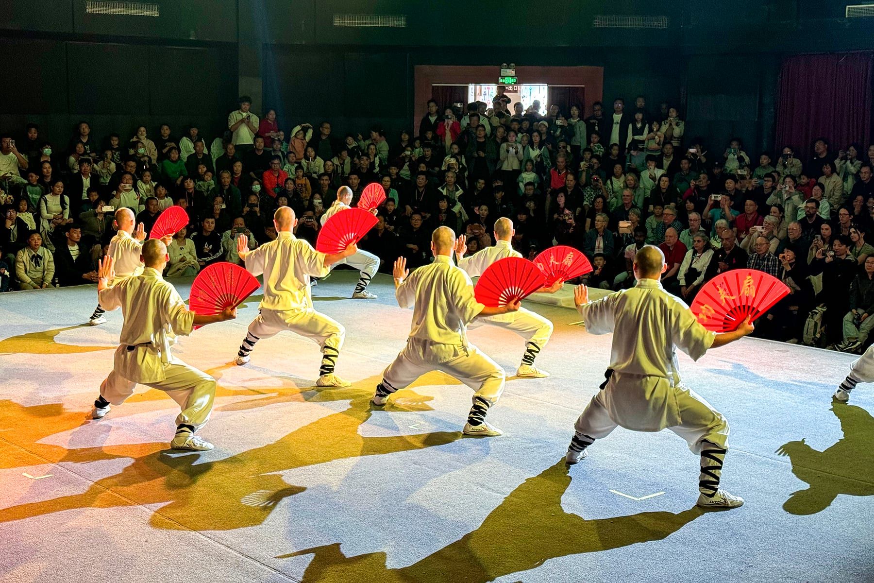 Shaolin-Vorführung: Kämpfer in gelben Gewändern mit roten Fächern in tiefen Ständen auf einer Bühne vor Publikum