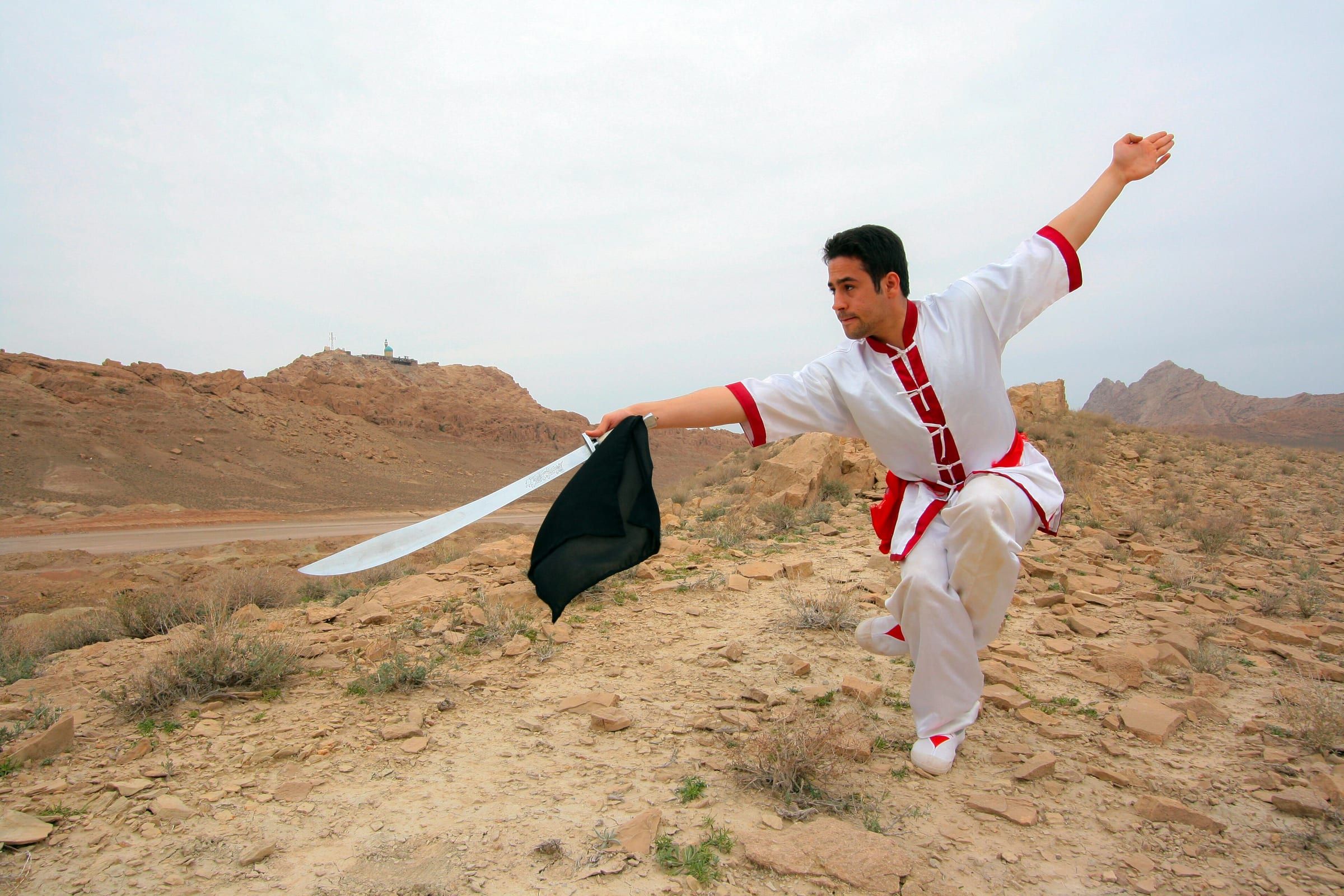 Wushu-Sportler in weiß-rotem Anzug mit Säbel (Dao) in einem weiten Ausfallschritt in hügeliger Landschaft