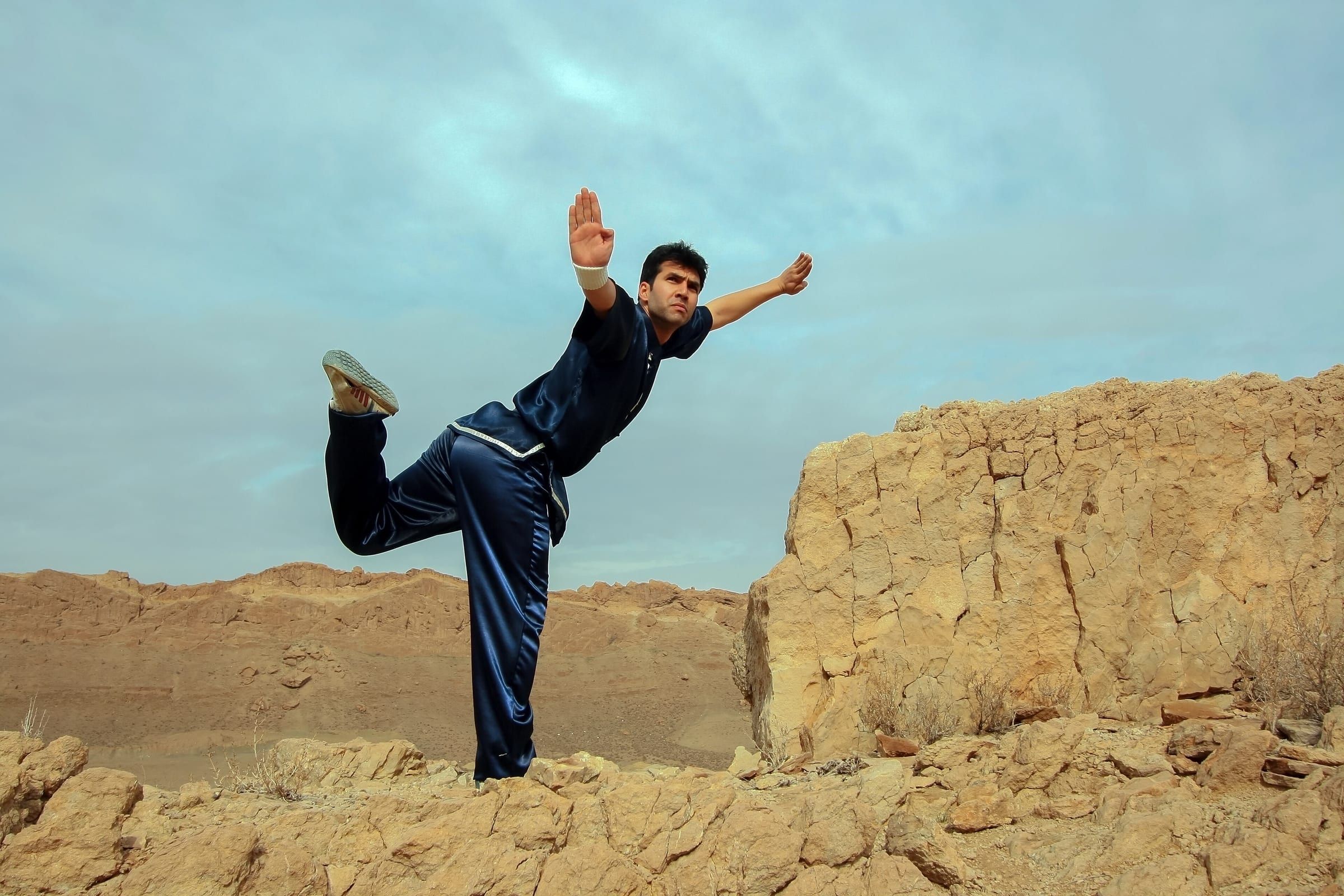 Kung-Fu-Sportler in dunkler Trainingskleidung in einem hohen Einbeinstand mit gestrecktem Bein auf einem Felsgrat, dahinter weiter Himmel